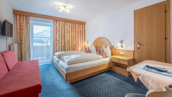 A cozy hotel room with a double bed and a red couch. There is a balcony with an outdoor view and wood-colored furniture. - Haus Timmelseck - Obergurgl 