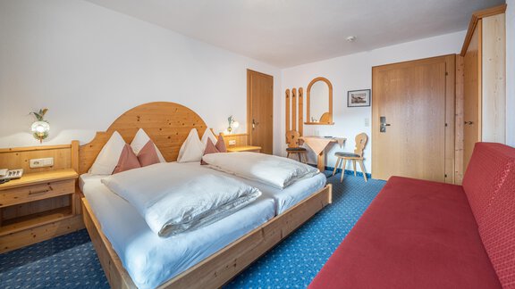 A cozy hotel room with a double bed, a red sofa, and wooden furniture. The space is bright and cheerfully designed. - Haus Timmelseck - Obergurgl 