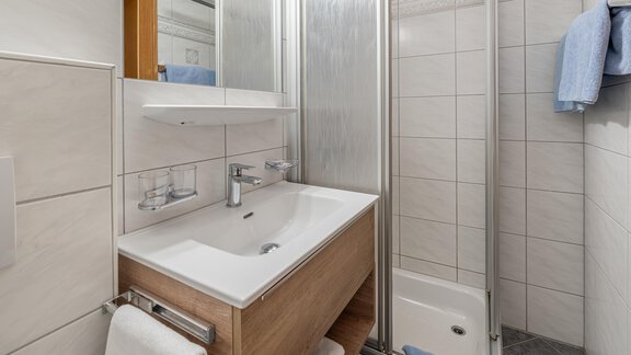 A modern bathroom with a shower, a sink, and wooden cabinets. The walls are lightly tiled and there are fresh towels in blue. - Haus Timmelseck - Obergurgl 