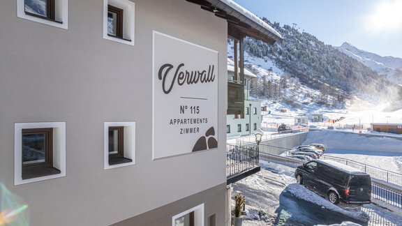 A modern building with a sign that shows "Verwall" and the number 115. Snow-capped mountains can be seen in the background. - Haus Verwall Appartements - Obergurgl 