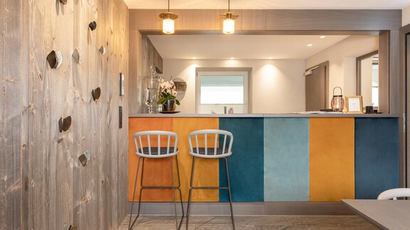 A modern kitchen with a colorful bar counter and two stools. The walls are partially decorated with wood and climbing grips. - Haus Verwall Appartements - Obergurgl 