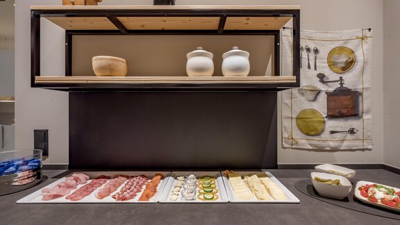 A stylish presentation of various cold cuts and cheese slices on a white plate. In the background, decorative containers and a wall hanging with kitchen utensils are visible. - Hotel-Garni Pirchhütt - Obergurgl 