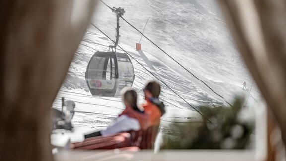 A cozy scene with two people sitting on a terrace and enjoying the view. In the background, a cable car and snowy landscape can be seen. - Hotel Gotthard-Zeit - Obergurgl 