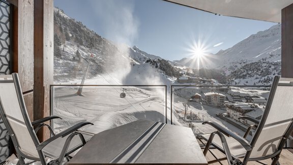 A terrace with chairs offers a view of a snowy mountain landscape. The sun is shining over the mountains and there is a ski lift station nearby. - Hotel Gotthard-Zeit - Obergurgl 