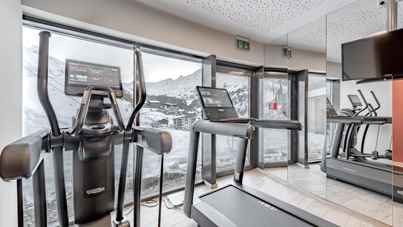 A modern fitness room with a view of the mountains. In front of a large window stand a treadmill and an elliptical trainer. - Hotel Gotthard-Zeit - Obergurgl 