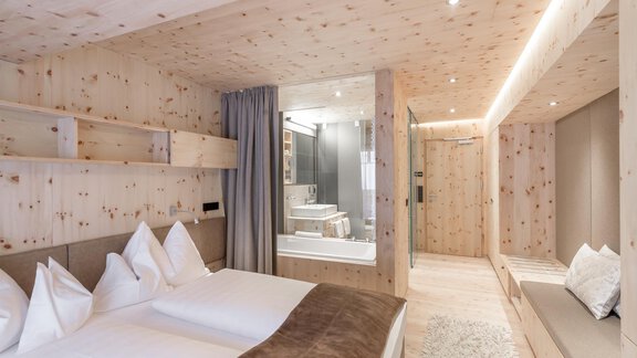 A modern room with wooden paneling and a cozy double bed. The bathroom is separated by a glass wall and feels bright and inviting. - Hotel Olympia - Obergurgl 