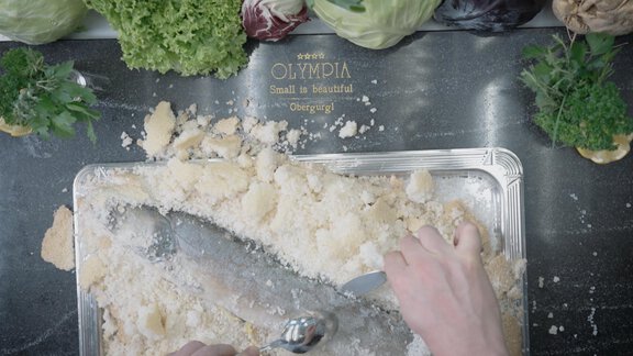 A kitchen table with fresh vegetables and crushed bread. A hand is cutting the ingredients on a silver tray. - Hotel Olympia - Obergurgl 