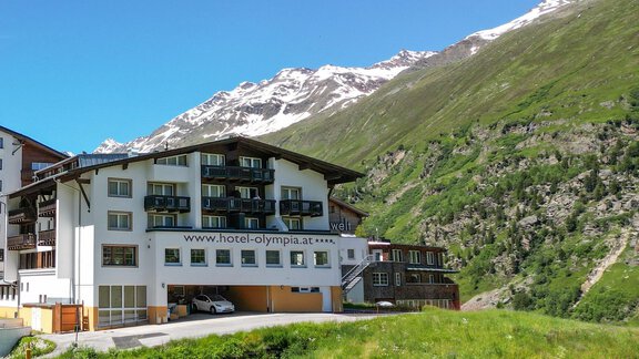 A modern hotel surrounded by green meadows and mountain landscapes. In the background, snow-capped peaks and a clear blue sky are visible. - Hotel Olympia - Obergurgl 