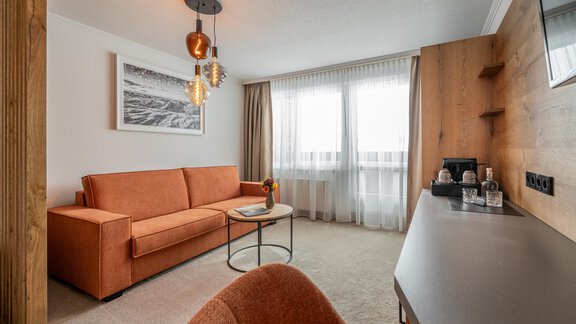 A modern living room with an orange sofa and a small table.  
Large windows provide plenty of light and a cozy atmosphere. - Hotel Olympia - Obergurgl 