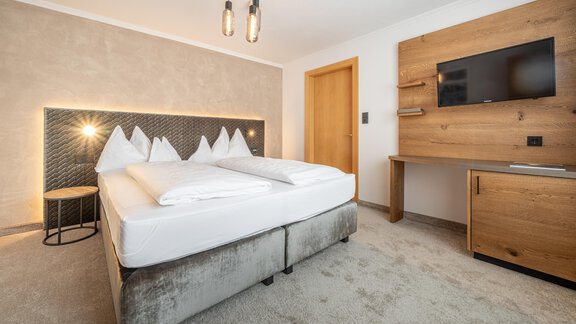 A modern bedroom with a large double bed and a television on the wall. The room features bright colors and wooden details, creating a cozy atmosphere. - Hotel Olympia - Obergurgl 