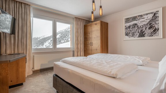 A modern bedroom with two beds and a large window that offers a view of the snow-covered mountains. The furnishings are cozy with wooden furniture and neutral colors. - Hotel Olympia - Obergurgl 