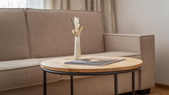 A modern living room with a beige sofa and a round wooden table. On the table, there is a vase with dried flowers and a magazine. - Hotel Olympia - Obergurgl 