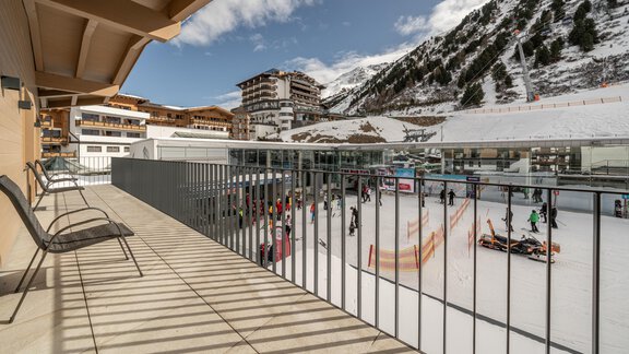 A balcony with chairs, overlooking a busy ice skating rink. In the background, snow-covered mountains and buildings are visible. - Mein Gaisberg - Obergurgl 