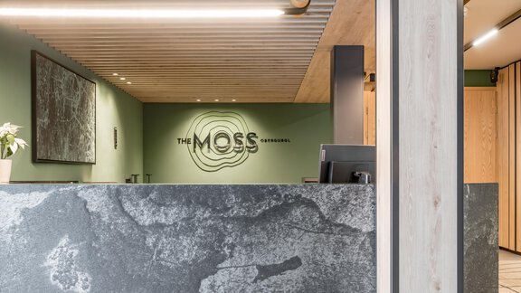 A modern reception area with a stylish gray tabletop and a relaxing green wall. The logo "THE MOSS" is prominently visible. - The Moss - Your unique hideaway - Obergurgl 