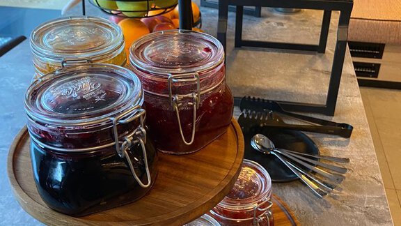 A buffet with various jams in jars on a wooden board. In the background, fresh fruit and pastries are present. - The Moss - Your unique hideaway - Obergurgl 