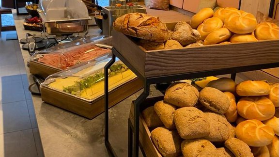 A rich breakfast buffet with various types of bread on wooden shelves. In the background, fresh fruits and other delicacies can be seen. - The Moss - Your unique hideaway - Obergurgl 