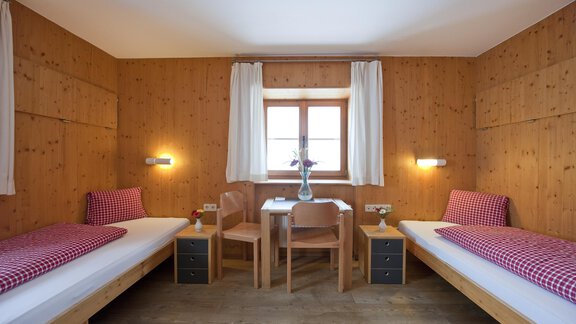 A cozy room with two single beds and wooden paneling. A table and two chairs are positioned in front of a window with a vase and flowers. - Universitätszentrum Obergurgl - Obergurgl 