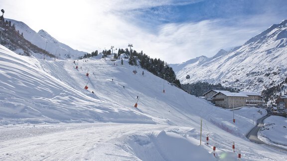 A snow-covered slope with skiers and a clear blue sky. In the background, mountains and a lodge are visible. - Universitätszentrum Obergurgl - Obergurgl 