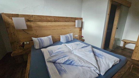 A cozy bedroom with a wooden bed and soft blue bedding. Two lamps are installed on the wall and there is a small seating area. - Almhof Mareil - Ochsengarten