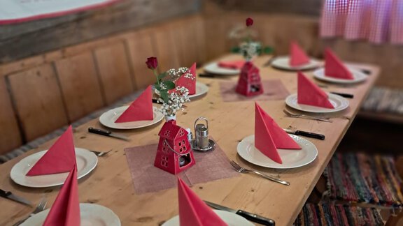 Ein rustikaler Tisch in einer gemütlichen Hütte ist liebevoll gedeckt. Es gibt rote Servietten und Blumen in der Tischmitte. - Almhof Mareil - Ochsengarten