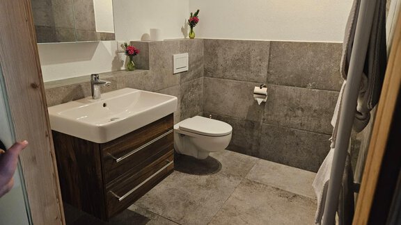Ein modernes Badezimmer mit einem Waschbecken und einer Toilette. Die Wände sind mit grauen Fliesen gestaltet und es gibt einige Blumen als Dekoration. - Almhof Mareil - Ochsengarten