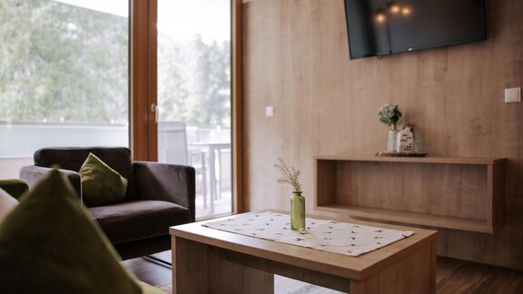 Ein modernes Wohnzimmer mit einem Holztisch und einem großen Fernseher an der Wand. Die Fenster lassen viel Tageslicht herein und bieten einen Blick ins Freie. - Apartment Waldesruh - Ochsengarten
