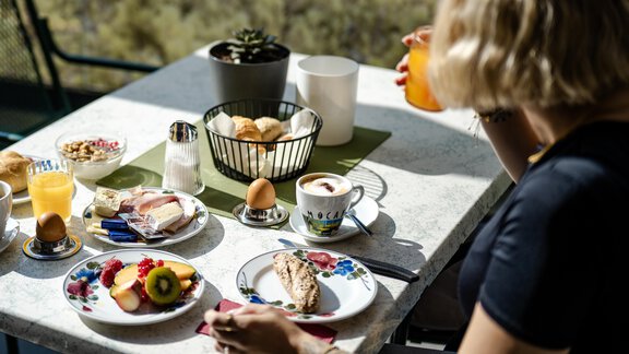 Ein schöner Frühstückstisch mit verschiedenen Speisen wie Obst, Käse, Brötchen und einem Getränk. Im Hintergrund ist eine Person zu sehen, die das Frühstück genießt. - Gasthof Waldesruh - Ochsengarten