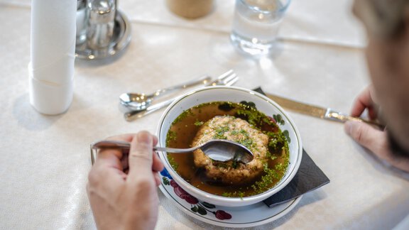 Eine Person isst aus einer Schüssel mit Brühe und einem gefüllten Knödel. Der Tisch ist schön gedeckt mit Besteck und einem Glas Wasser. - Gasthof Waldesruh - Ochsengarten