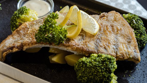 Ein gegrillter Fischfilet mit Zitrone und frischen Brokkoliröschen. Daneben befindet sich ein kleiner Becher mit Dip. - Gasthof Waldesruh - Ochsengarten