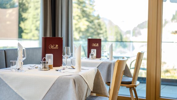 Ein eleganter Restauranttisch mit weißen Tischdecken und rotbraunen Menüs. Große Fenster bieten einen schönen Blick auf die natürliche Umgebung. - Gasthof Waldesruh - Ochsengarten