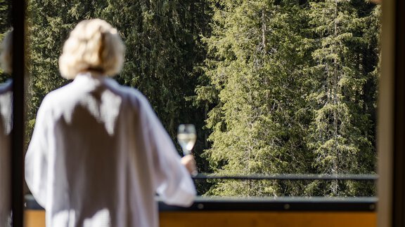 Eine entspannte Person in einem Bademantel steht auf einem Balkon und hält ein Glas in der Hand. Im Hintergrund sind grüne Bäume und die Natur zu sehen. - Gasthof Waldesruh - Ochsengarten