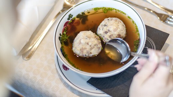 Eine Person isst eine Schüssel mit Brühe und zwei Knödeln. Der Tisch ist gedeckt mit Besteck und einem weißen Tischtuch. - Gasthof Waldesruh - Ochsengarten