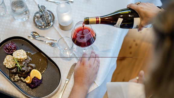 Ein elegant gedeckter Tisch mit einem Teller, der feine Speisen präsentiert. Eine Person gießt Wein in ein Glas. - Gasthof Waldesruh - Ochsengarten