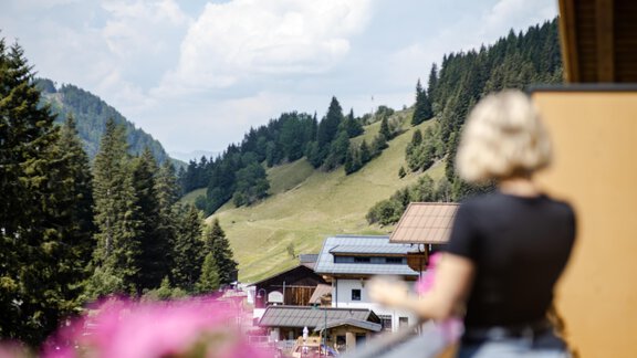 Eine wunderschöne Berglandschaft mit grünen Hügeln und bewaldeten Flächen. Im Vordergrund steht eine Person auf einem Balkon mit pinken Blumen. - Gasthof Waldesruh - Ochsengarten