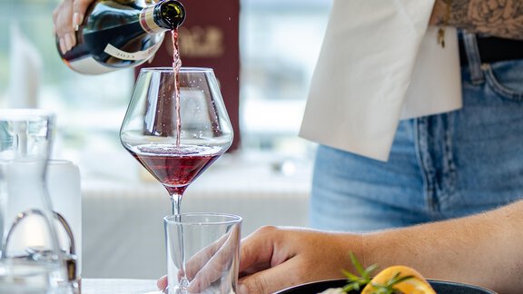 Ein Kellner schenkt Wein in ein Glas ein, während eine Mahlzeit auf dem Tisch steht. Auf dem Teller sind verschiedene Speisen und eine Garnitur zu sehen. - Gasthof Waldesruh - Ochsengarten
