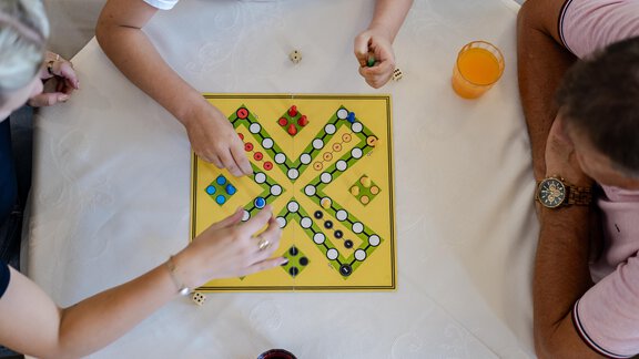 Zwei Personen spielen ein Brettspiel auf einem Tisch. Auf der Tischdecke liegen Würfel und ein Glas mit Getränk. - Gasthof Waldesruh - Ochsengarten