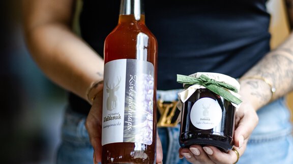 Eine Person hält eine Flasche Getränk und ein Glas Marmelade. Beide Produkte haben ansprechende Etiketten und sind in der Hand einer Person zu sehen. - Gasthof Waldesruh - Ochsengarten