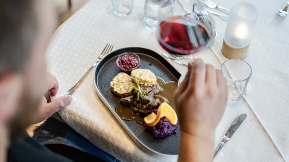 Ein köstlicher Teller mit verschiedenen Beilagen und Fleisch, serviert auf einem schwarzen Teller. Eine Weinglas wird hervorgehoben, während der Gast das Essen genießt. - Gasthof Waldesruh - Ochsengarten