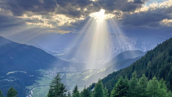An impressive mountain landscape with sun rays breaking through the clouds. Green meadows and coniferous trees characterize the scenery. - Acherberg Alm - Oetz