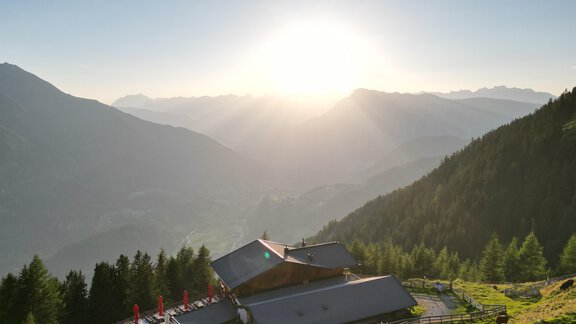 A mountain landscape with a beautiful house surrounded by trees. The sun rises above the mountains, creating a picturesque atmosphere. - Acherberg Alm - Oetz