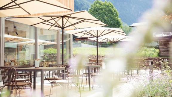 An inviting outdoor café with tables and sun umbrellas. In the background, green hills and trees can be seen. - All-Suite Resort Ötztal - Oetz