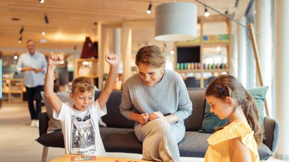 A cheerful family is playing together at a table. The children are showing excitement while a woman watches them. - All-Suite Resort Ötztal - Oetz