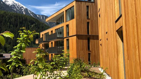 Modern wooden buildings surrounded by lush vegetation. In the background, the mountains can be seen, providing a picturesque backdrop. - All-Suite Resort Ötztal - Oetz