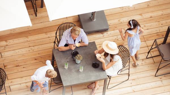 A family sits at a table on a wooden terrace and enjoys food. Children are playing and moving around the table. - All-Suite Resort Ötztal - Oetz