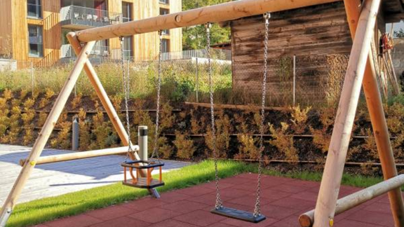 A playground with a swing in a green environment. In the background, mountains and modern residential buildings are visible. - All-Suite Resort Ötztal - Oetz