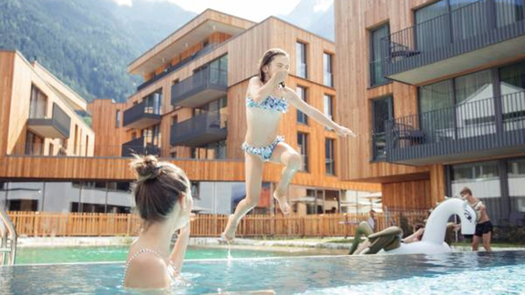 A girl joyfully jumps into a pool while another person sits in the water watching. In the background, modern wooden structures and a mountainous landscape can be seen. - All-Suite Resort Ötztal - Oetz