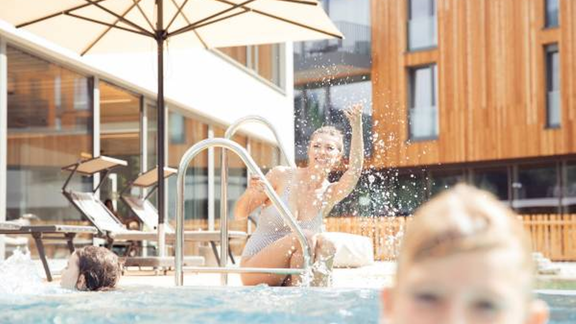 A woman is laughing and splashing water at the pool. In the background, there are loungers and a modern building visible. - All-Suite Resort Ötztal - Oetz