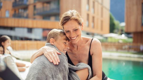 A happy woman is hugging a boy at the edge of a swimming pool. In the background, modern buildings and green landscape can be seen. - All-Suite Resort Ötztal - Oetz