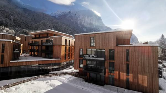Modern wooden buildings in a snow-covered landscape with mountains in the background. The sun is shining brightly, creating a cozy atmosphere. - All-Suite Resort Ötztal - Oetz