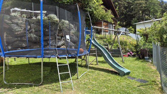 Ein Garten mit einem Trampolin und einer Rutsche. Im Hintergrund sind ein Haus und einige Bäume zu sehen. - Alpenlove - Oetz
